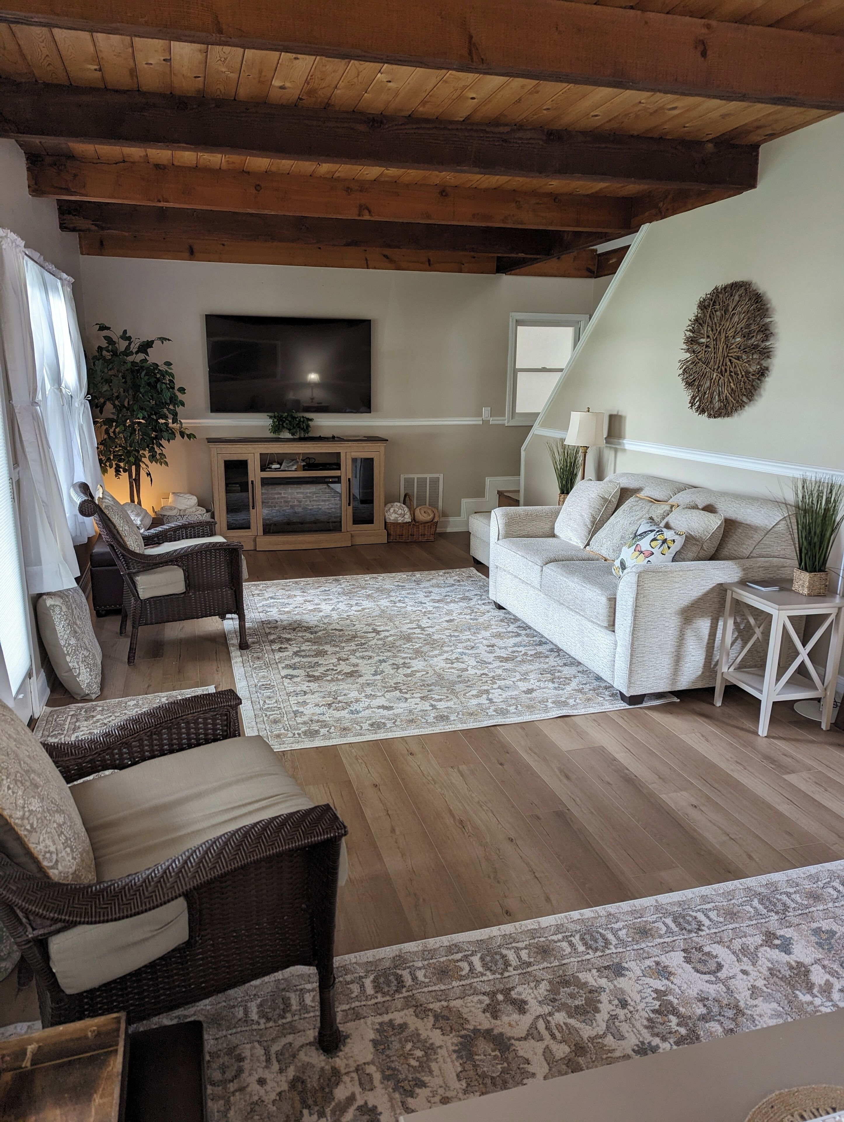 A cozy living room with a wooden ceiling, a large flat-screen TV, a white sofa, a wicker chair, and a patterned rug.