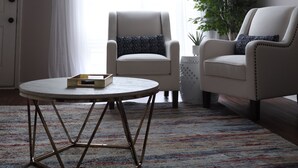 Una sala de estar con una mesa redonda, dos sillones, una planta en maceta y un ventanal con cortinas blancas.