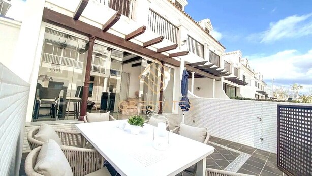 A patio area with wicker furniture and a white table, overlooking a white building with balconies and a clear sky.
