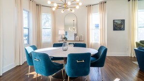 Een eetruimte met een ronde tafel, zes blauwe stoelen, een kroonluchter en een grote spiegel aan de muur.
