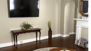 Una sala de estar con televisor de pantalla plana, una mesa de centro de madera, un tapete redondo, un ventilador de techo y una chimenea.