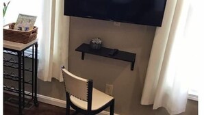 Uma sala de estar com televisão de tela plana na parede, uma cadeira de madeira, uma prateleira pequena abaixo da TV e um sapateiro à esquerda.