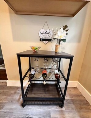 A black metal table with a wine rack, a bowl of snacks, and a welcome sign on the wall behind it.