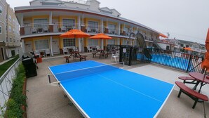 A pool table on a deck with a pool in the background, surrounded by outdoor seating and a building with multiple balconies. - Sea Chest Motel  (Wildwood Crest)