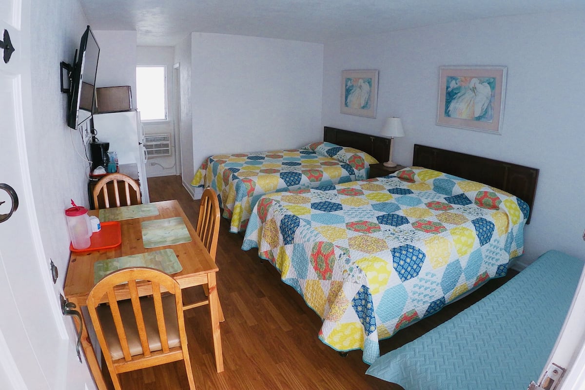Une chambre avec deux lits simples, une table en bois avec des chaises, un téléviseur fixé au mur et une fenêtre.