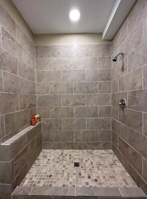 Eine Dusche mit grauen Fliesen, ein Ablagebereich mit Toilettenartikeln und ein gefliester Boden.