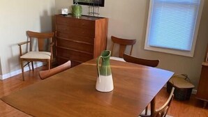 Une salle à manger avec une table en bois et six chaises, un vase vert posé sur la table et une fenêtre avec des stores.