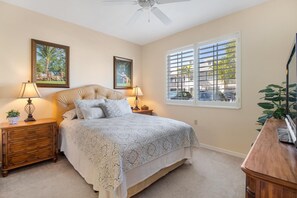 A bedroom with a bed, two nightstands, a ceiling fan, a window with blinds, and two framed pictures on the wall. - Luxury Naples Golf Condo (Naples)