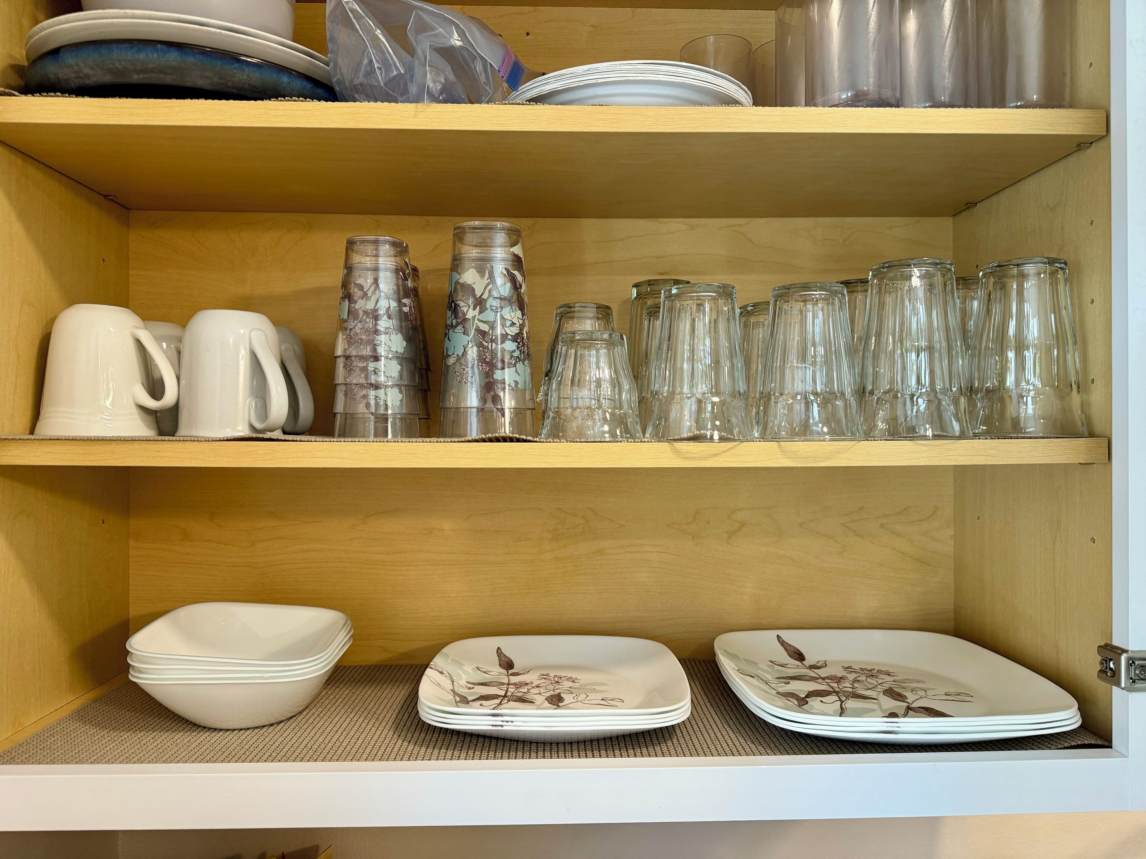 A wooden shelf with white and floral-patterned dishes, clear glass mugs, and a plastic bag.