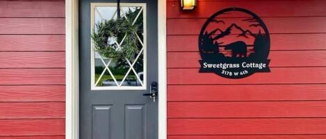 A red building with a gray door, a window with a plant, and a logo for Sweetgrass Cottage at 3178 W 4th.