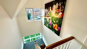 Una escalera con barandilla de madera y balaustradas blancas, que lleva a un rellano con un cuadro floral grande y una ventana con vistas a un césped verde.