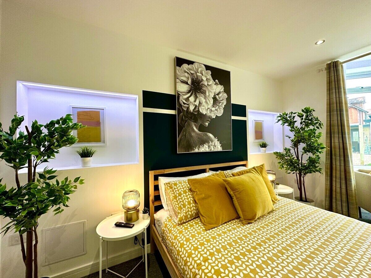 A modern hotel room with a large bed, a bedside table with a lamp, a wall-mounted artwork, and two potted plants.