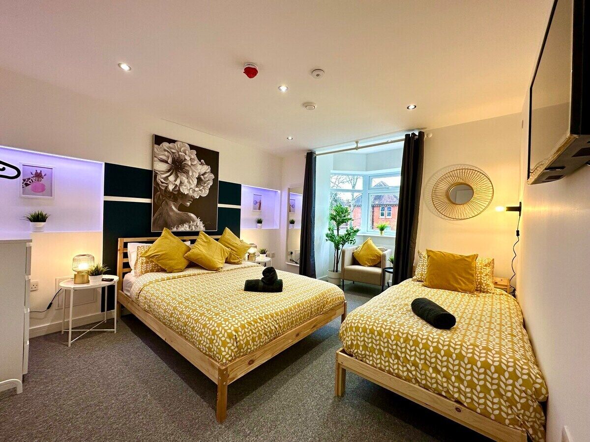 A hotel room with two beds, each with a patterned bedspread and yellow pillows. There is a small table with a lamp, a chair, and a wall-mounted TV. The room has a modern design with a large window and a view of the outside.