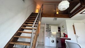 A staircase with wooden steps and metal railings leading to a kitchen area with a washing machine, a dining table, and a chair. - Charming cocoon (Cabourg)