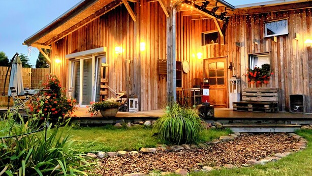 A rustic wooden barn-style building with a porch, a small garden, and string lights. - 50sqm apartment with two terraces, barbecue area and large outdoor area (Putbus)