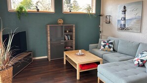 A living room with a grey sofa, a wooden coffee table, a TV on a stand, a bookshelf, and two windows with plants. - 50sqm apartment with two terraces, barbecue area and large outdoor area (Putbus)