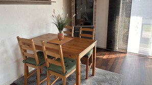 A dining area with a wooden table, four chairs with green cushions, a vase with plants, and a wooden shelf above the table. - 50sqm apartment with two terraces, barbecue area and large outdoor area (Putbus)