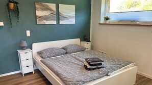 A neatly made bed with geometric patterned bedding, a bedside table with a lamp, and a wall-mounted shelf with a plant. - 50sqm apartment with two terraces, barbecue area and large outdoor area (Putbus)