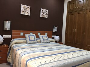 A bedroom with a bed, two wall-mounted light fixtures, a wooden wardrobe, and two wall art pieces. - Medina Azahara (Córdoba)