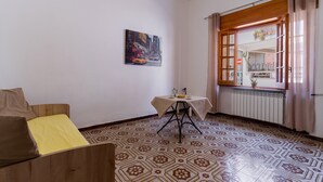 A room with a patterned floor, a wooden sofa with yellow cushions, a small table with a white tablecloth, and a window with curtains.
