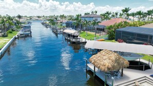 A canal with boats, a thatched-roof structure, and a modern building with a red roof. - Intervillas Florida - Bella Villa 141 (Cape Coral)