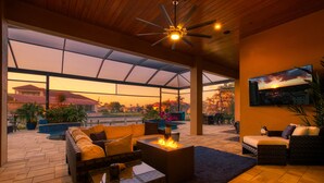 A spacious outdoor living area with a fire pit, comfortable seating, and a large TV screen displaying a sunset view. - Intervillas Florida - Bella Villa 141 (Cape Coral)