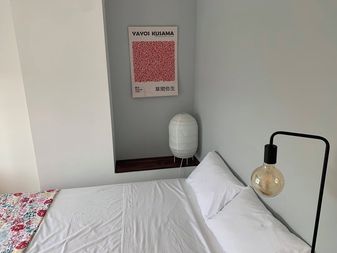 A neatly made bed with white linens, a wall-mounted light fixture, and a framed poster on the wall.