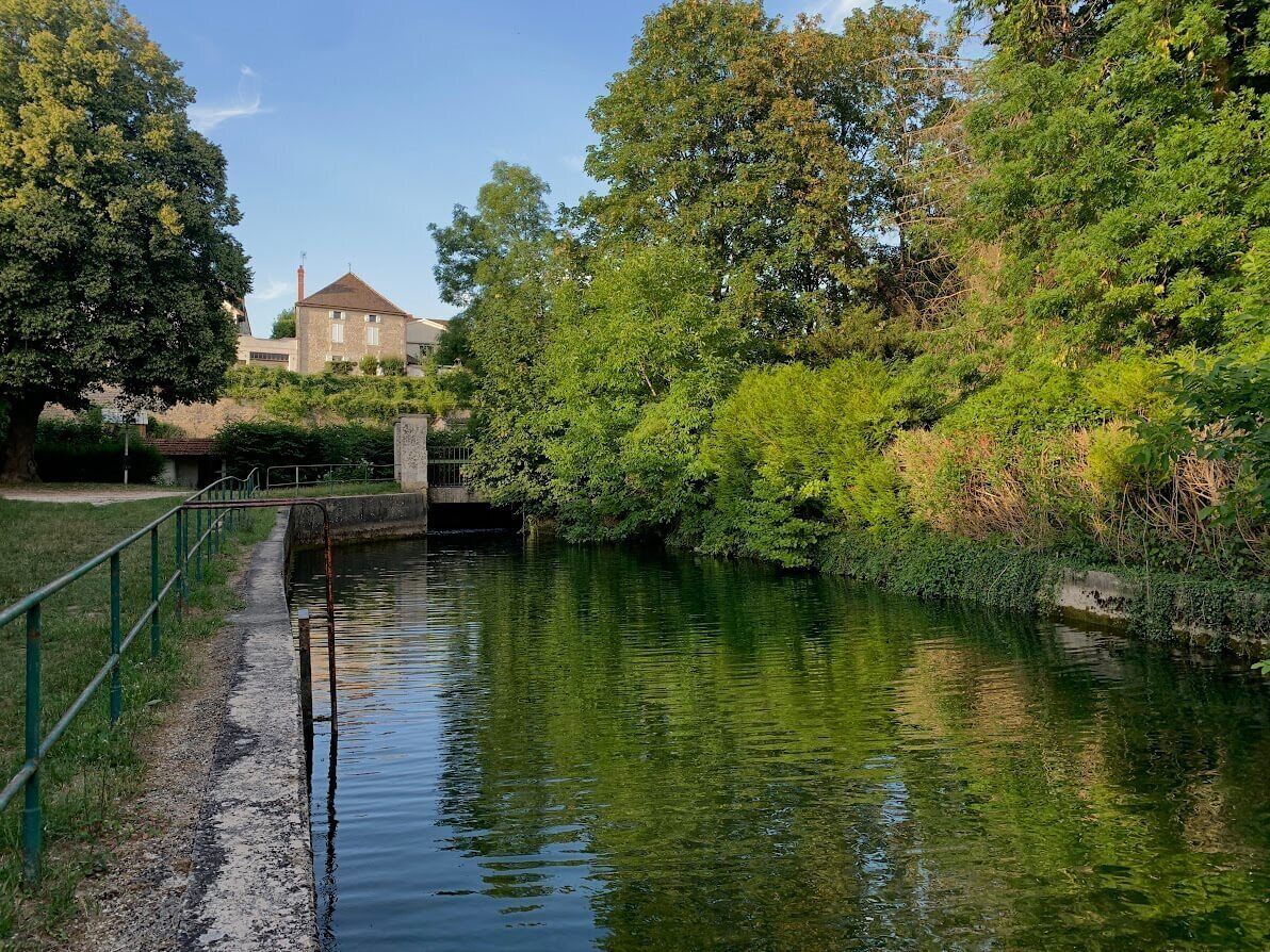 A calm river with a bridge, surrounded by lush greenery and a building in the background.