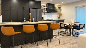 A modern kitchen with dark cabinets, a central island with bar stools, and a dining area with a table and chairs. - Off-Broadway • Boho Modern 3BR • King Suite • Huge Yard + Treehouse • Near River & Downtown (Boise)