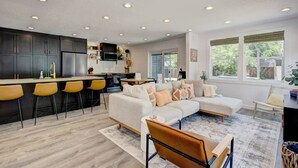 A modern kitchen with dark cabinets and a bar area, a living room with a sectional sofa and a coffee table, and large windows with a view of greenery. - Off-Broadway • Boho Modern 3BR • King Suite • Huge Yard + Treehouse • Near River & Downtown (Boise)