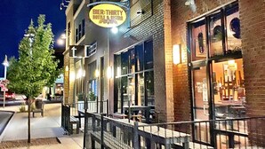 A building with a sign for 'Bier-THIRTY Bottle & Bistro' and a sidewalk with a tree and benches. - Off-Broadway • Boho Modern 3BR • King Suite • Huge Yard + Treehouse • Near River & Downtown (Boise)