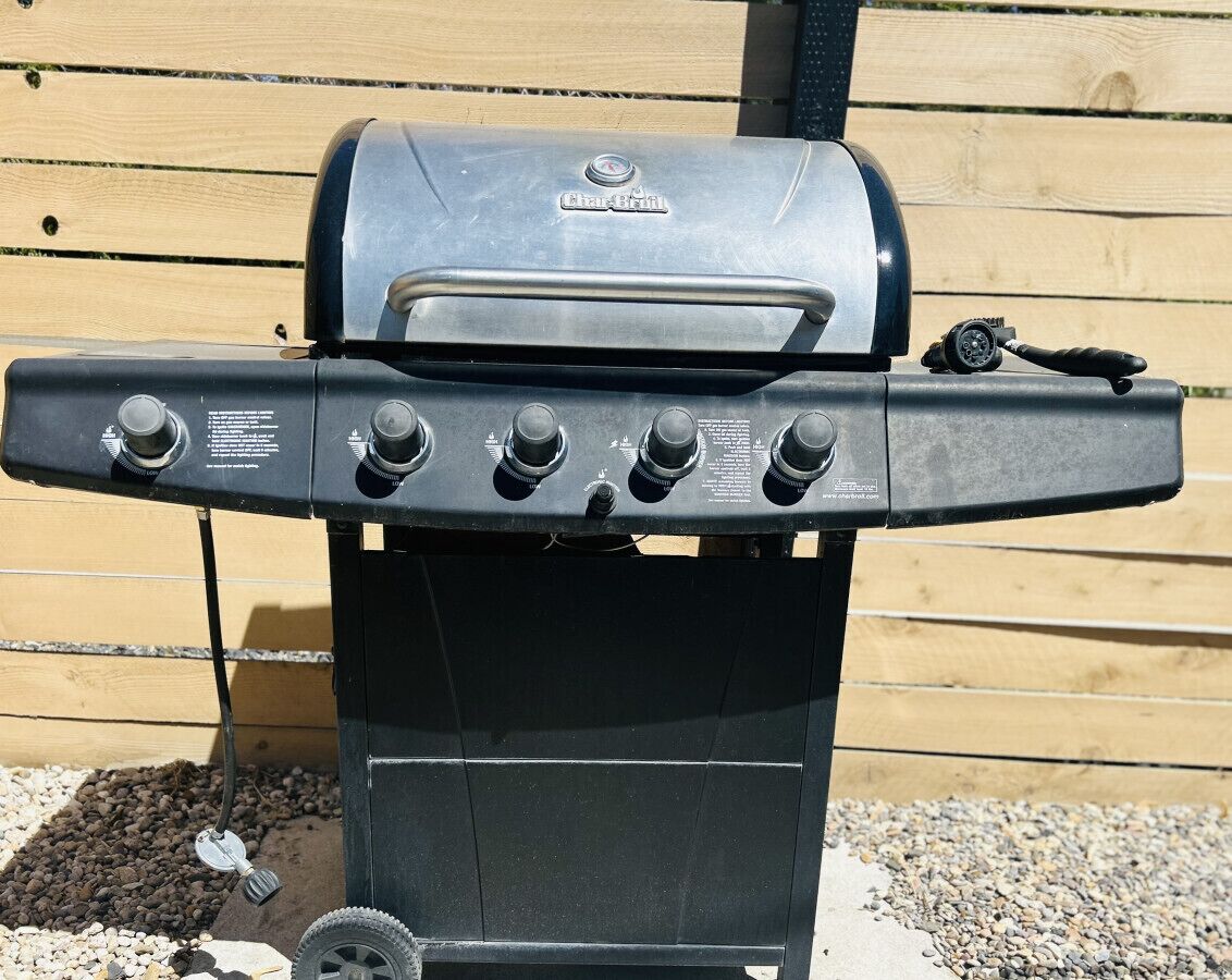 A Char-Broil gas grill with four burners and a side shelf, set against a wooden fence.