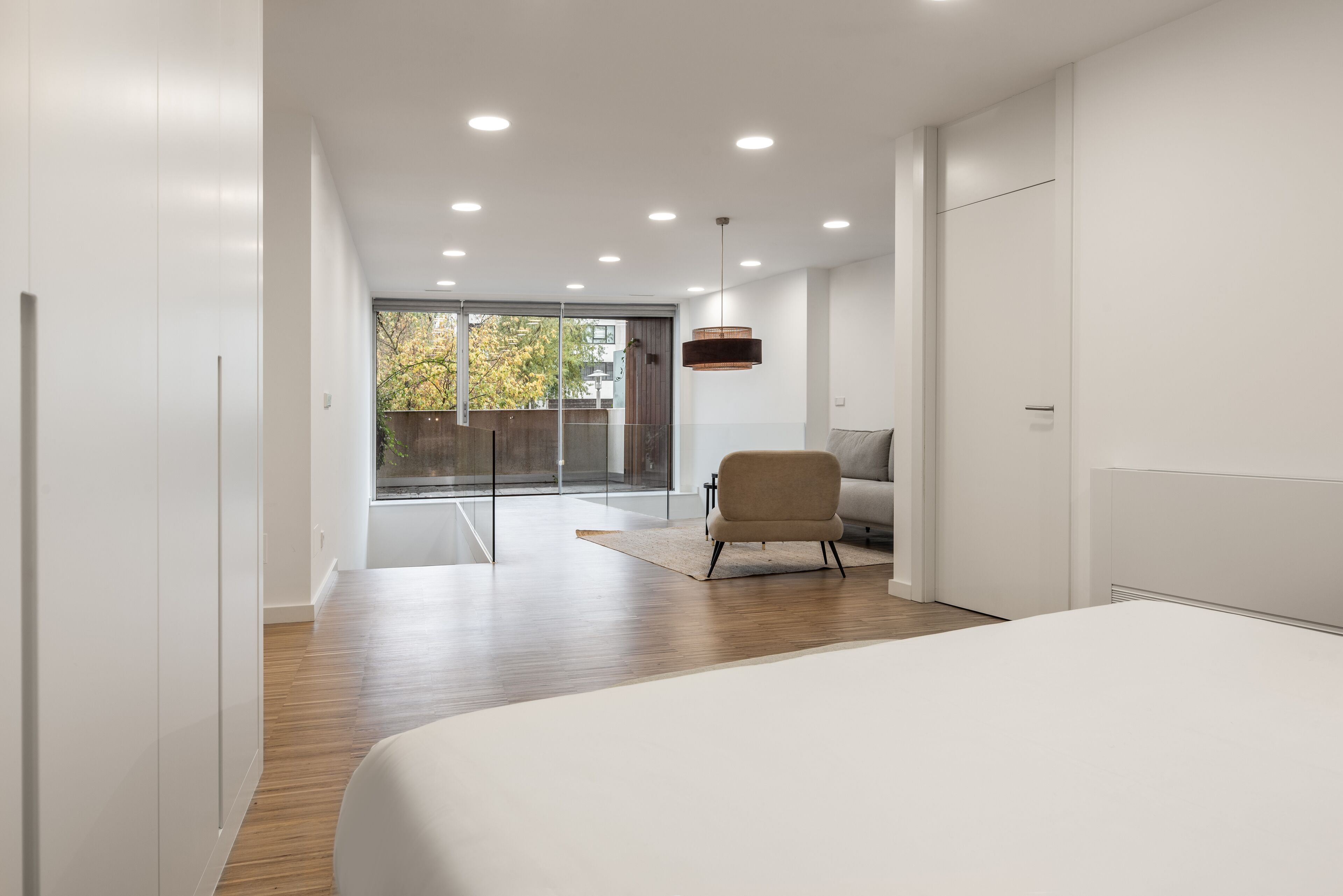A modern bedroom with a large bed, a sofa, and a chair. There is a sliding glass door leading to a balcony with a view of trees.
