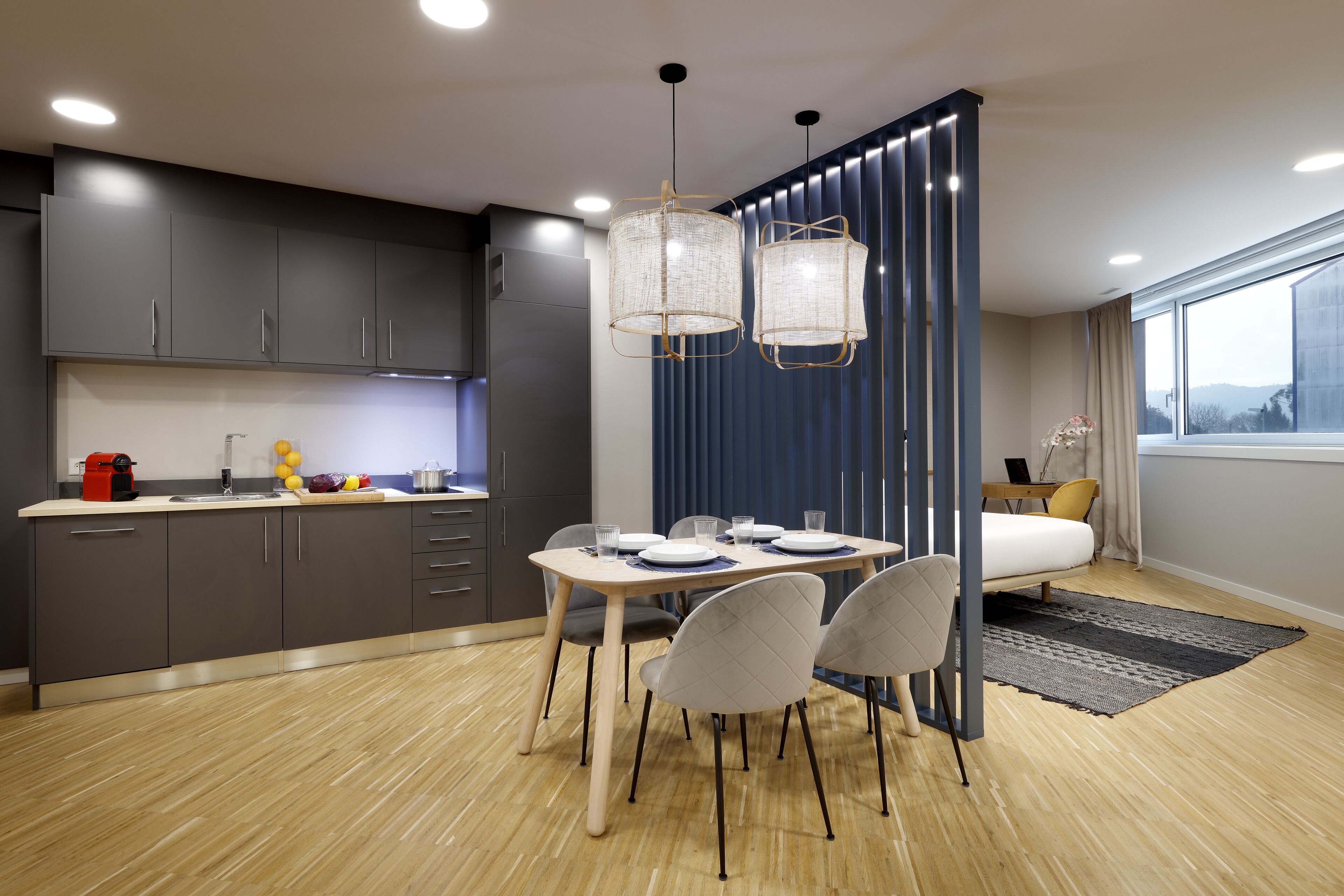 Une cuisine moderne avec des meubles de couleur sombre, une table à manger avec des chaises et des luminaires suspendus.