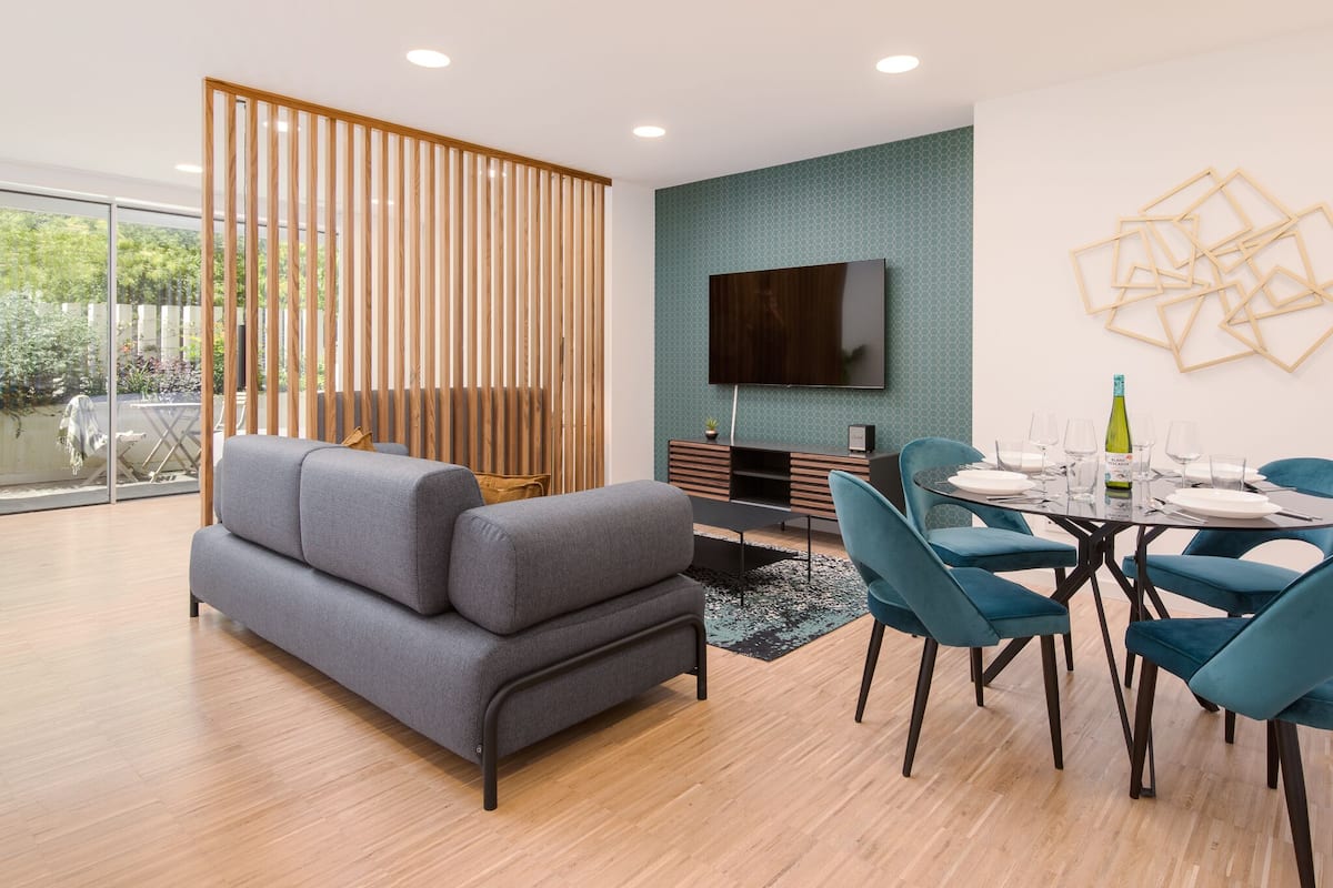 A modern living room with a grey sofa, teal chairs, a dining table with glasses and a bottle, a TV on the wall, and a wooden partition.