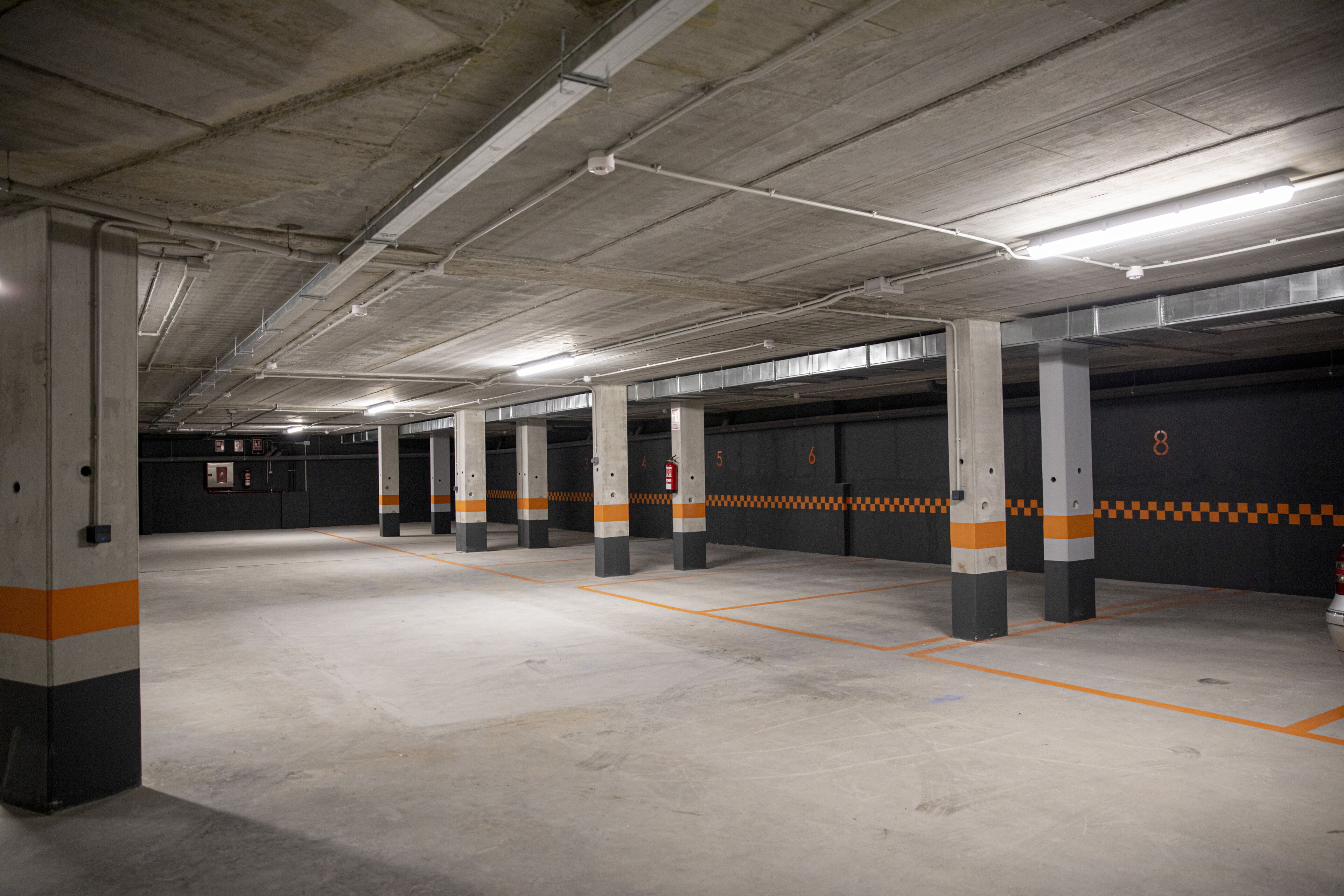 Un parking à plusieurs niveaux, avec des piliers en béton, des rayures orange et grises, et des places de stationnement numérotées.