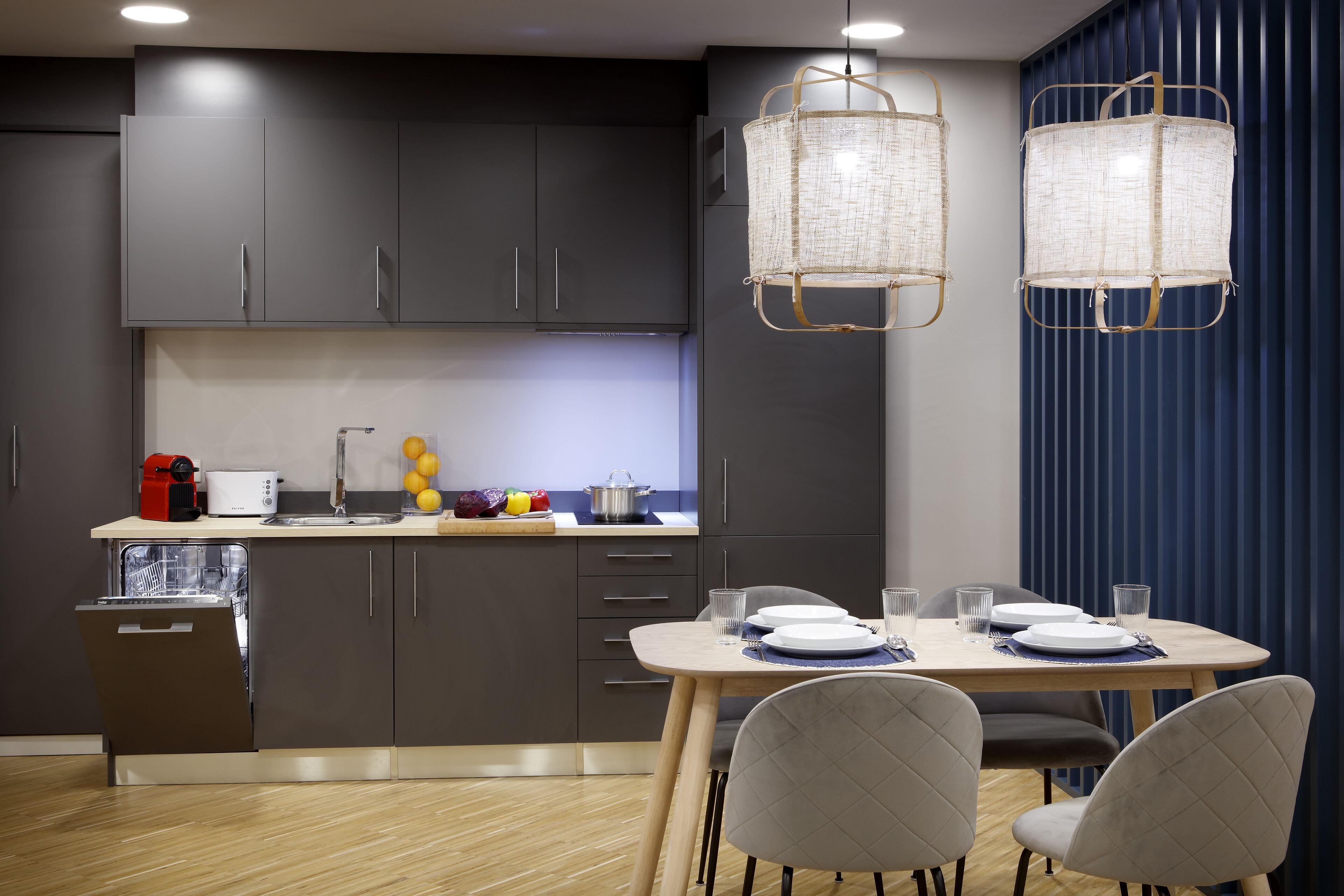 A modern kitchen with dark cabinets, a dishwasher, a toaster, and a fruit bowl. There is a dining table with chairs and a blue curtain in the background.