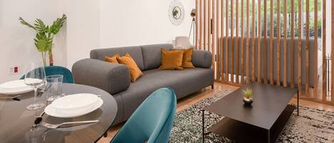 Una sala moderna con un sofá gris, una mesa de comedor dispuesta con platos y vasos, una silla azul y una pequeña planta en maceta sobre una mesa de centro negra.
