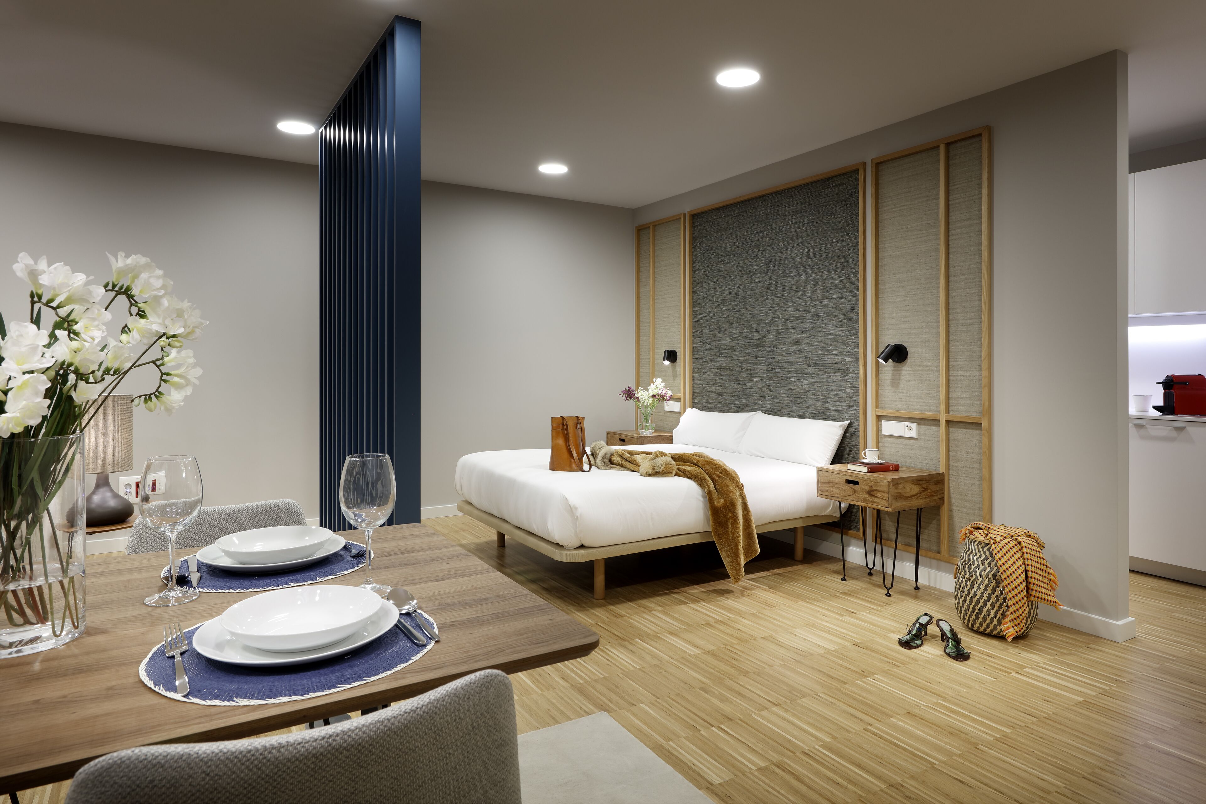 Una habitación de hotel moderna con piso de madera, una cama con ropa blanca, una mesita de noche con lámpara, una pequeña mesita auxiliar de madera, y un jarrón con flores blancas.