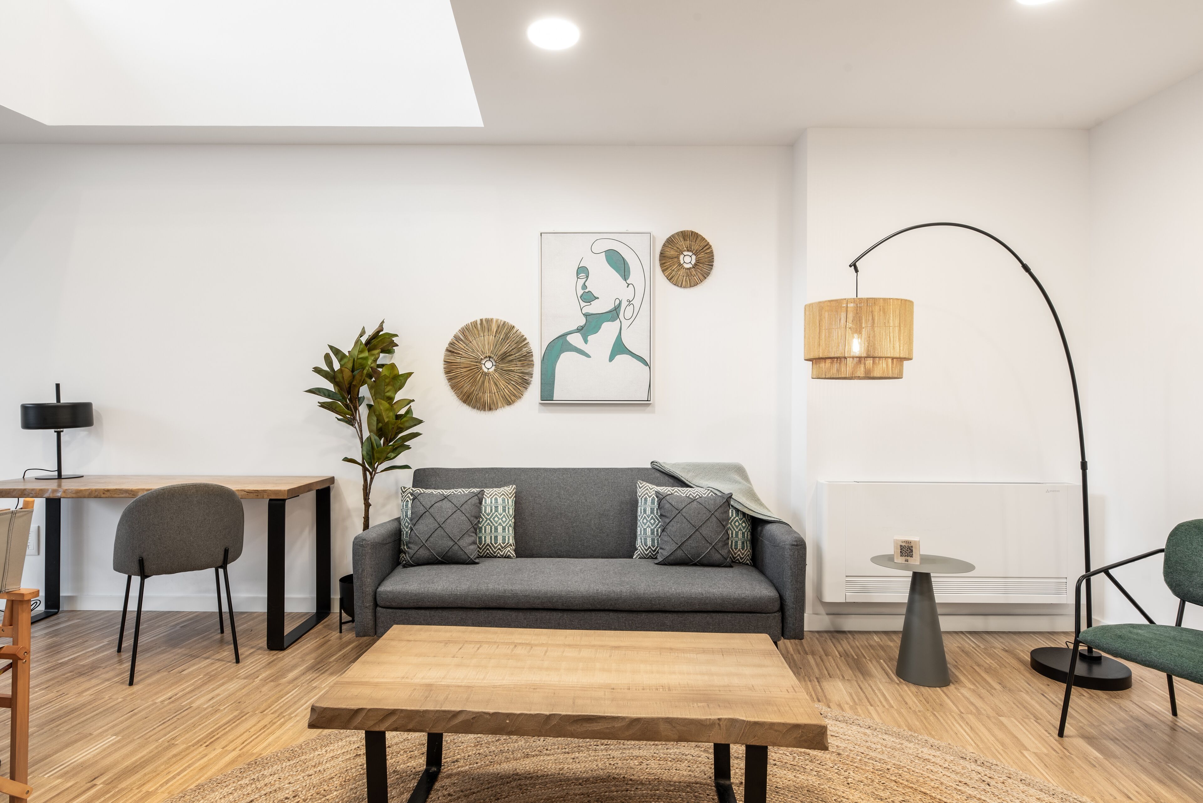 Ein modernes Wohnzimmer mit grauem Sofa, einem Holztisch, einem Schreibtisch mit Stuhl und einer Stehlampe.