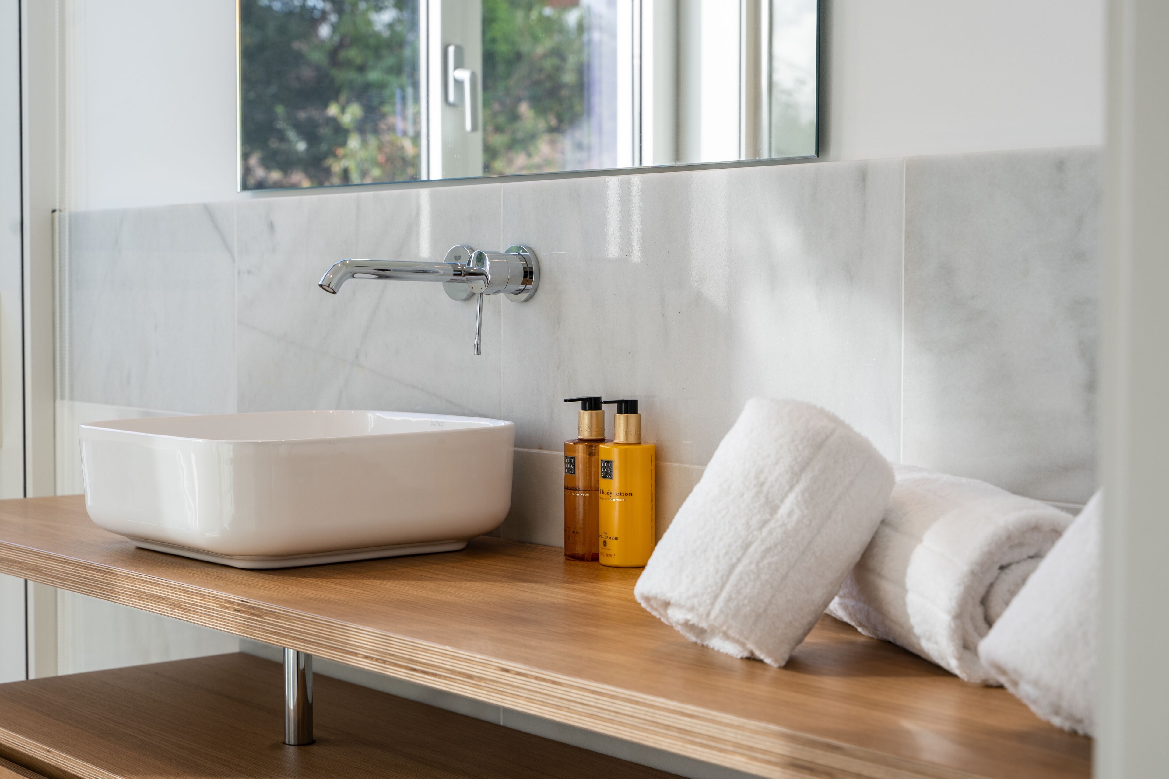 Une salle de bain avec un lavabo blanc, un miroir au-dessus et un plan de travail en bois. On y trouve deux flacons de lotion et une serviette enroulée sur le plan de travail.