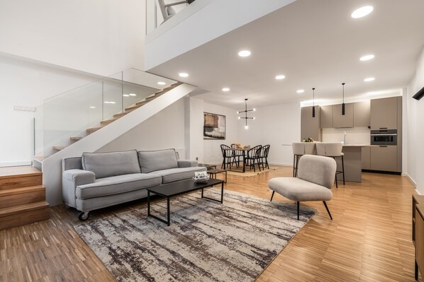Una sala de estar moderna con un sofá gris, una mesa de centro y un comedor con mesa y sillas. Una escalera conduce a un nivel superior.