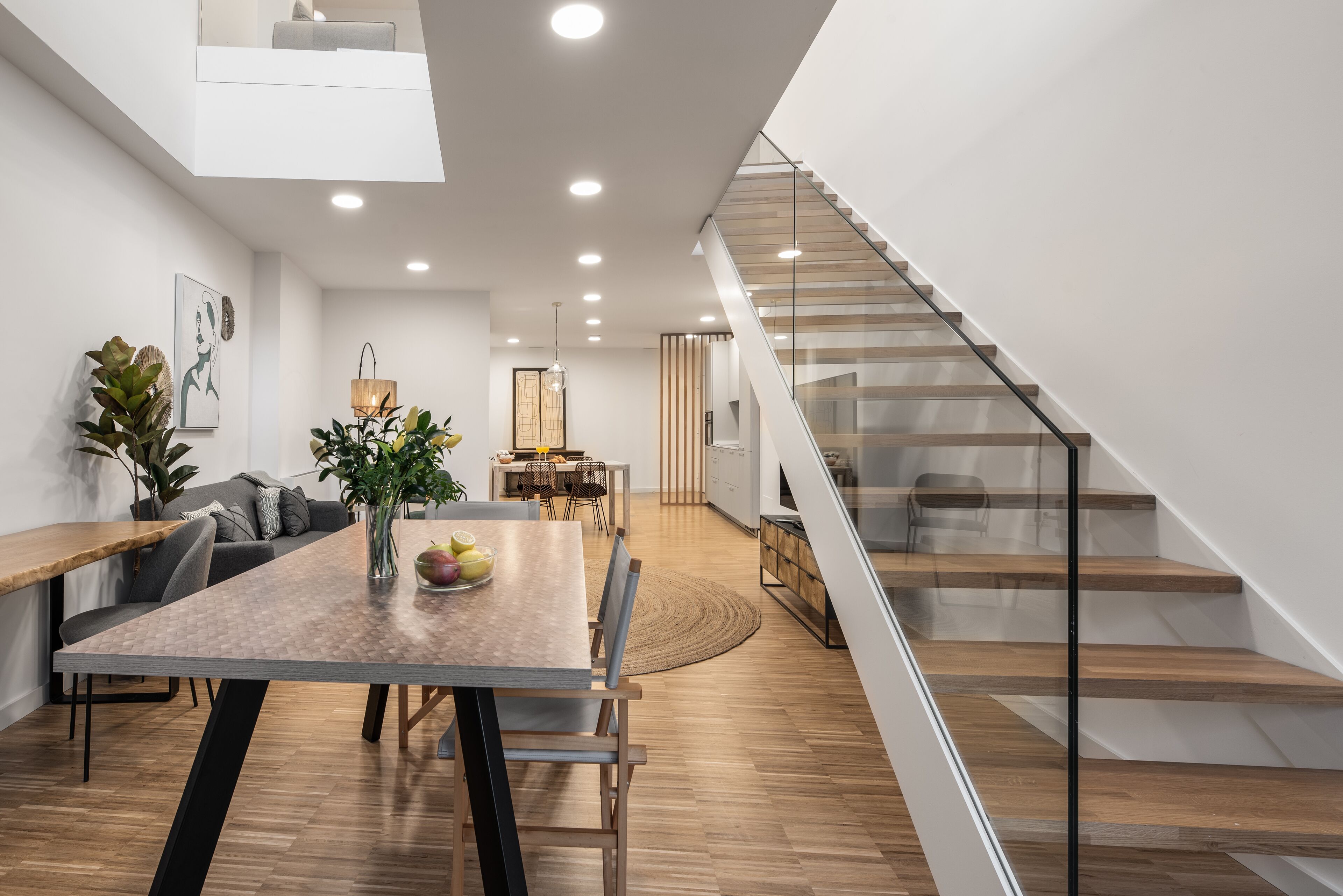 Un comedor moderno con una mesa y sillas de madera, un sofá y una escalera con barandillas de vidrio.