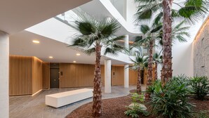 A modern lobby with a central seating area, palm trees, and a high ceiling with skylights.