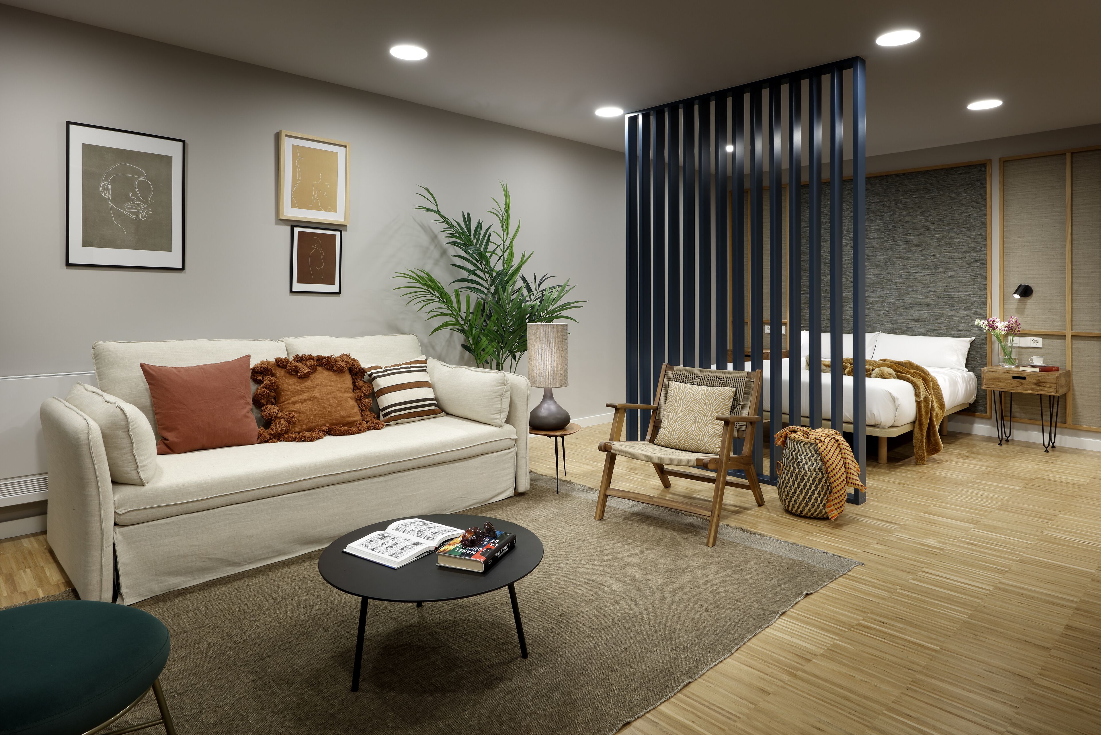 Una sala de estar moderna con sofá, sillón y mesa de centro. Se observa una planta, una lámpara y una cama al fondo.