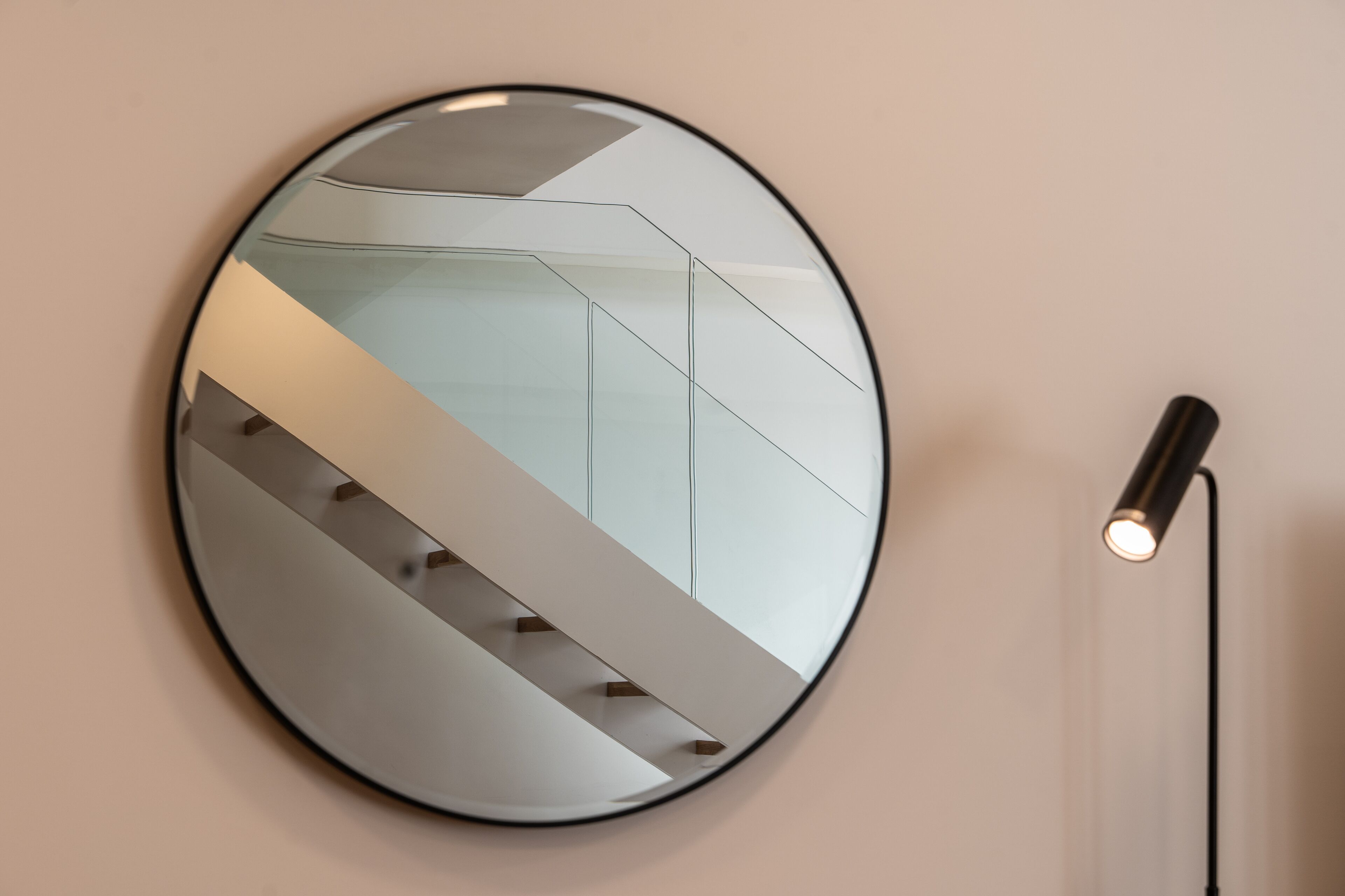 Un espejo redondo con un reflejo geométrico sobre una pared lisa, junto a una lámpara de pie.