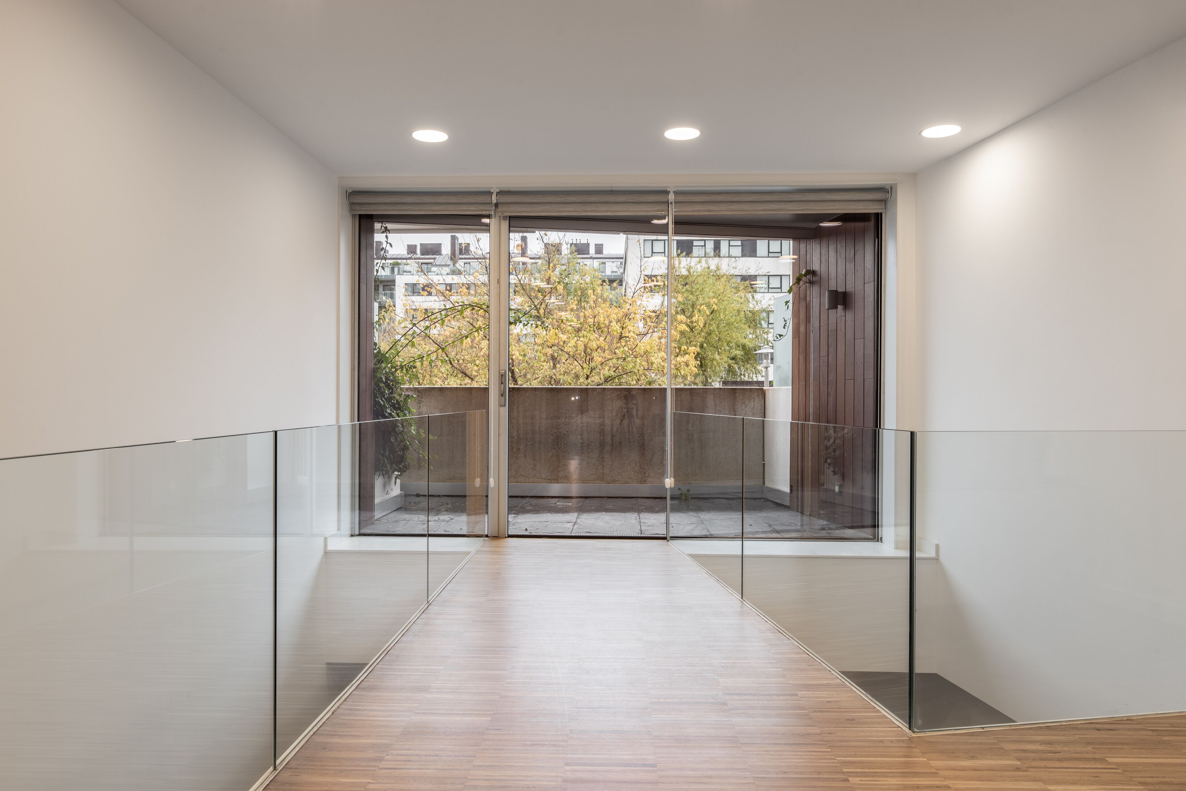 A modern interior with a glass railing, wooden flooring, and a view of trees and buildings outside.