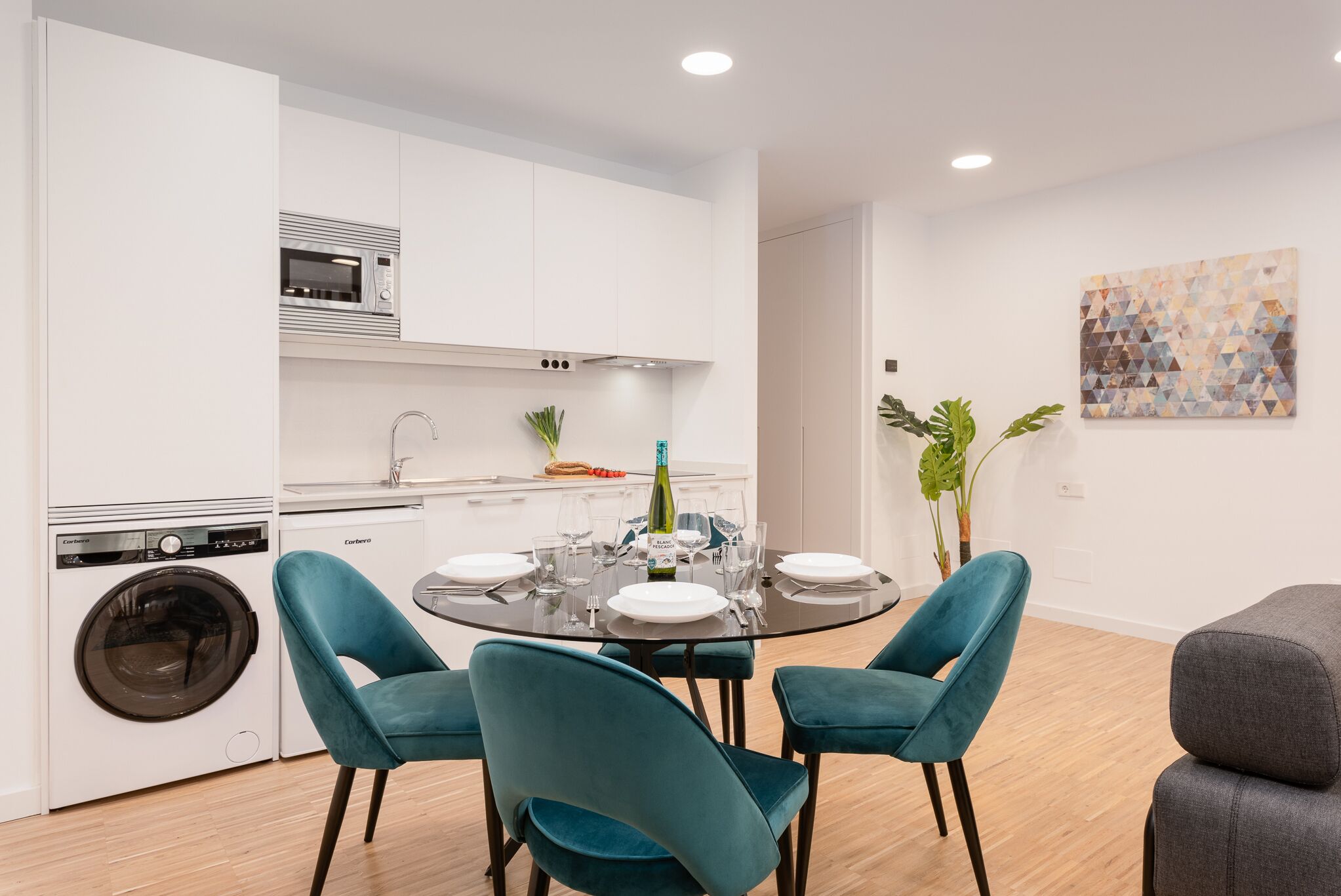 A modern kitchen with white cabinets, a built-in microwave, and a washing machine. There is a dining table with teal chairs and a bottle of wine.