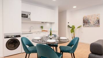 A modern kitchen with white cabinets, a built-in microwave, and a washing machine. There is a dining table with teal chairs and a bottle of wine.
