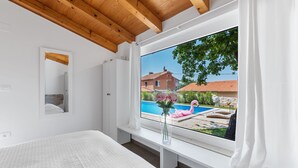 A bedroom with a large window, wooden ceiling beams, a bed, a bedside table, and a view of a swimming pool with a flamingo float. - Lavova in Brtonigla With 3 Bedrooms and 2 Bathrooms (Brtonigla)